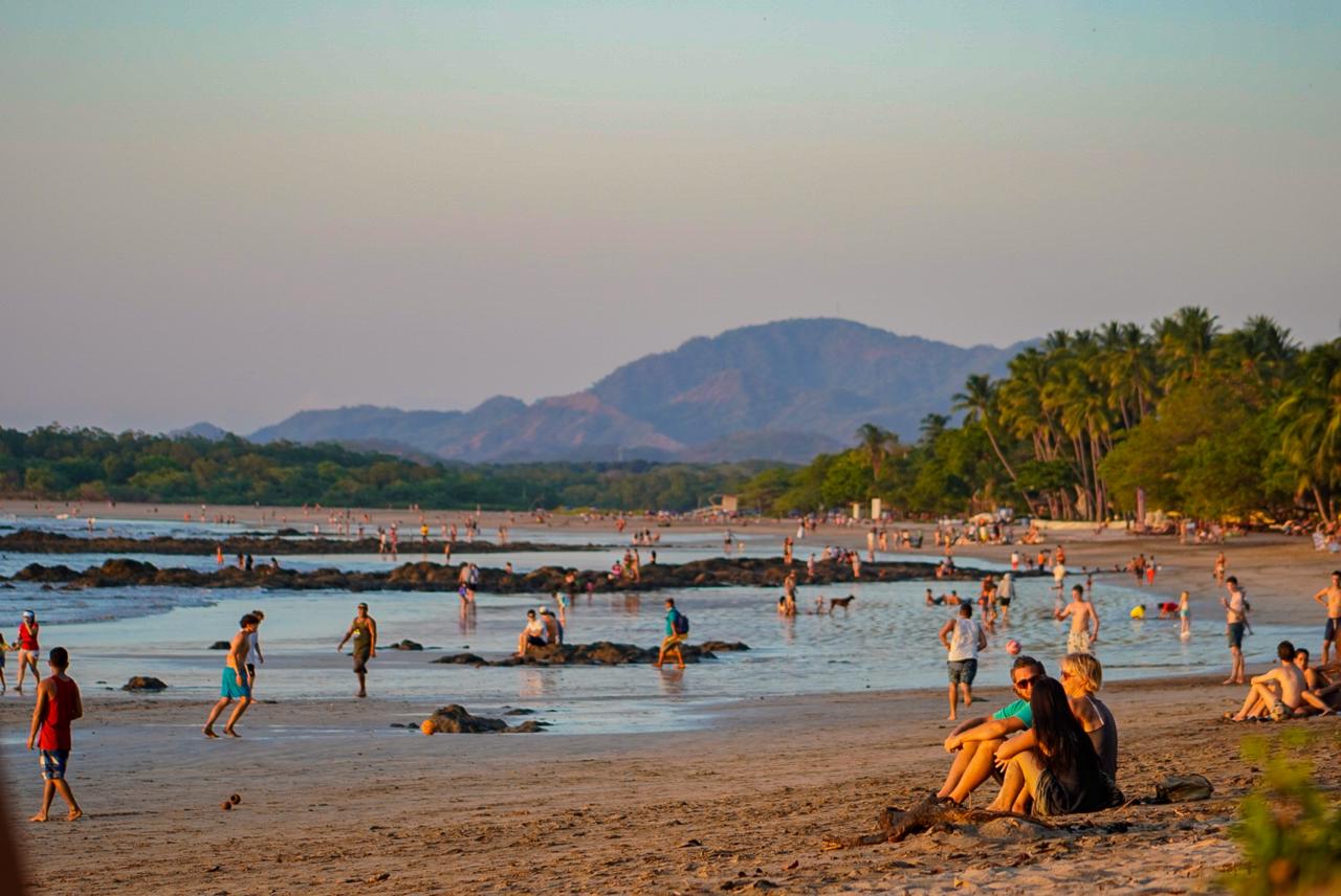 People enjoying a broad sandy beach with palm trees and mountains in the distance